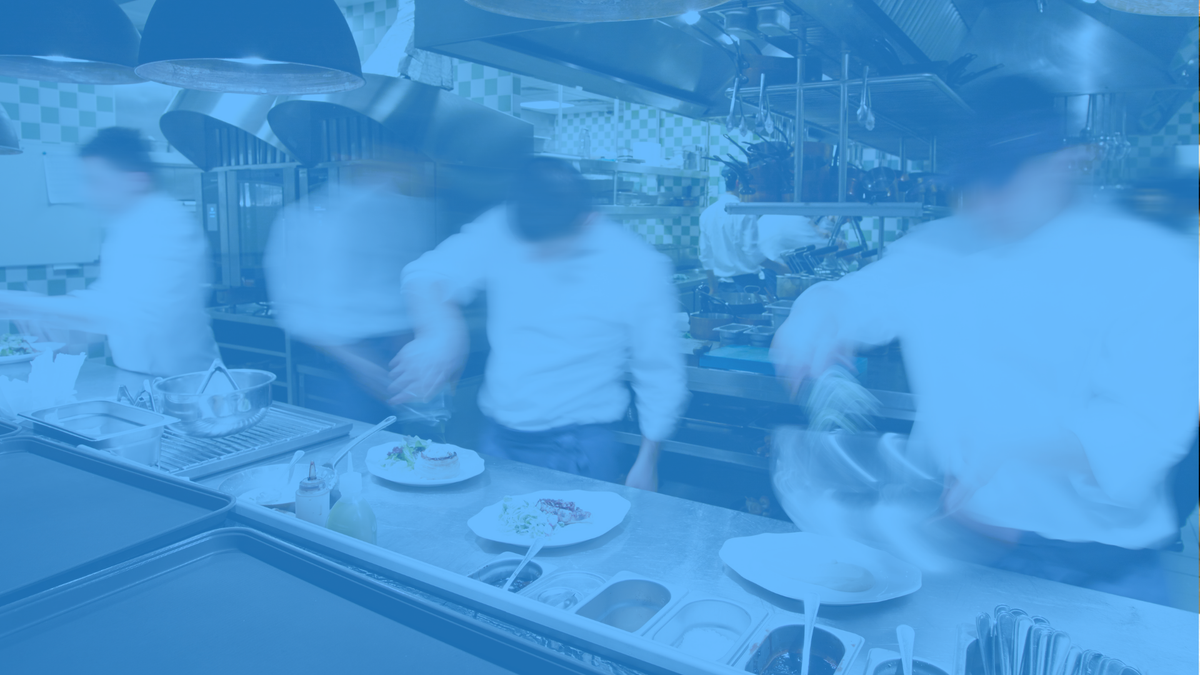 A blue-tinted photograph of a commercial kitchen with several blurred figures of chefs working behind a prep counter with plates and food items.